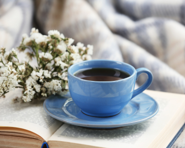 Une tasse à café et une soucoupe bleues surmontant un livre ouvert, avec des fleurs blanches à côté.