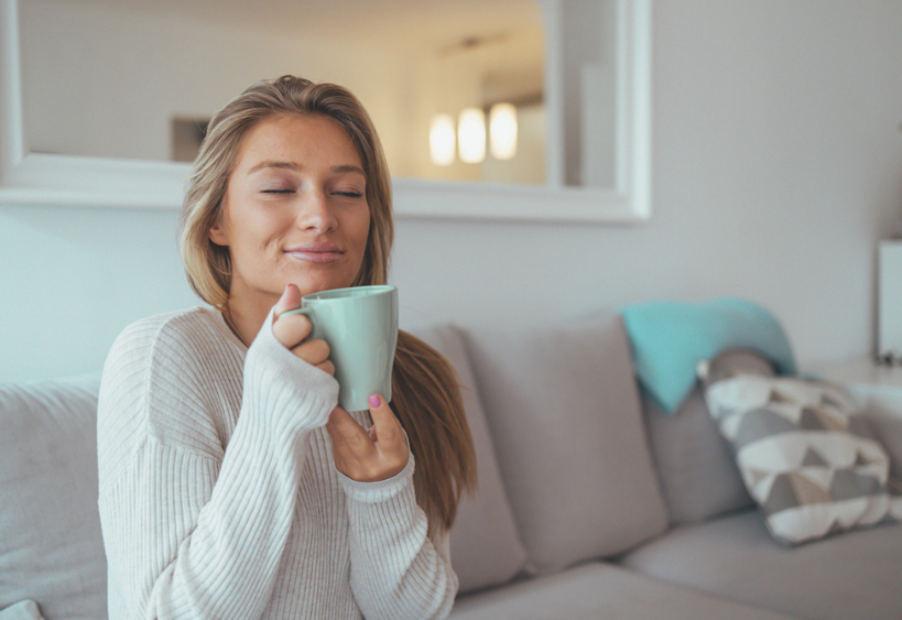 Une femme calme et satisfaite est assise sur un canapé gris, tenant une tasse couleur menthe et savourant une boisson chaude dans un salon lumineux et chaleureux.