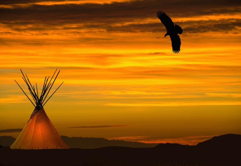 Tipi illuminé avec un aigle au‑dessus — coucher de soleil orangé