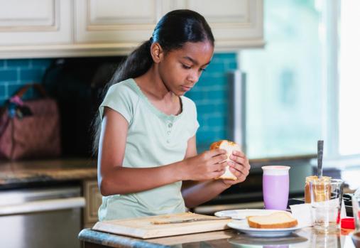 Une fillette métissée de 10 ans à la maison, assise au comptoir de la cuisine, sur le point de manger un sandwich au beurre d’arachide qu’elle vient de préparer