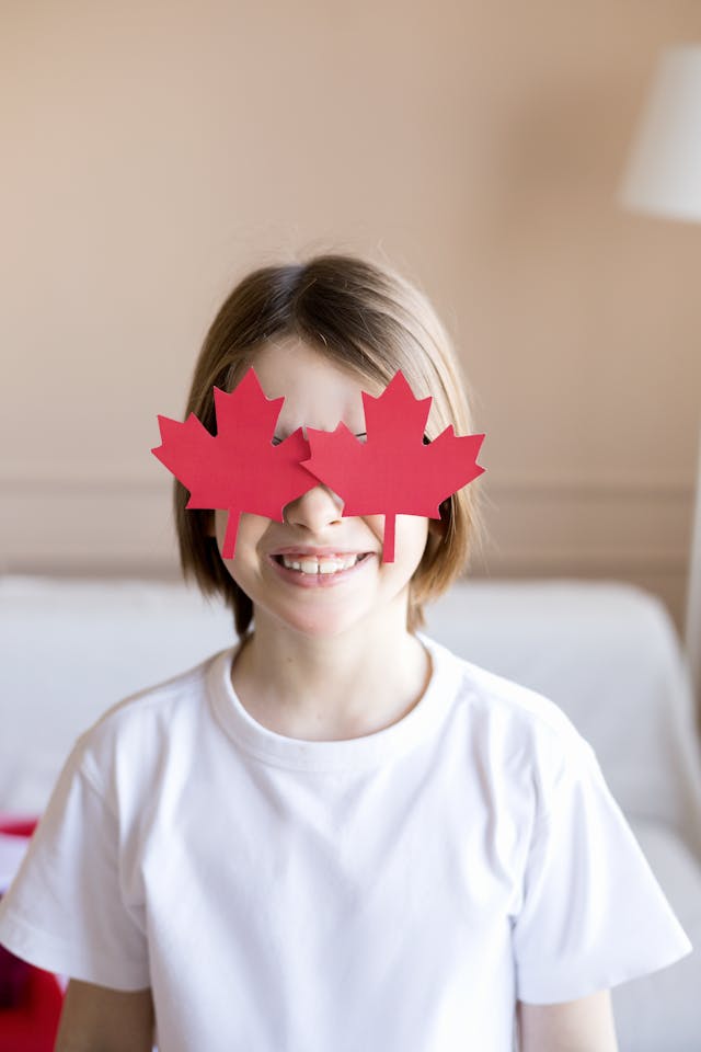 Un enfant souriant, portant des lunettes en forme de feuille d'érable, célèbre la fête nationale du Canada à l'intérieur.