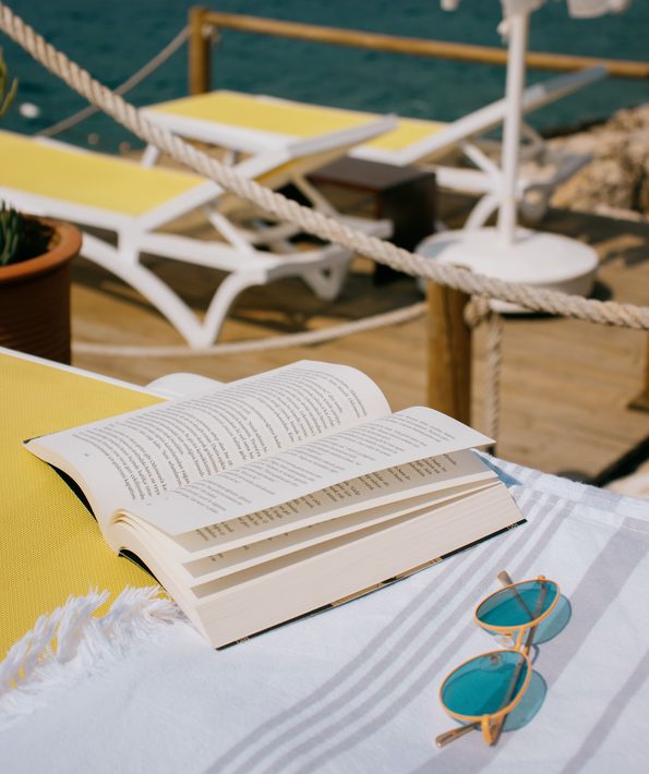 un livre posé sur un banc, à côté d’une serviette et de lunettes de soleil