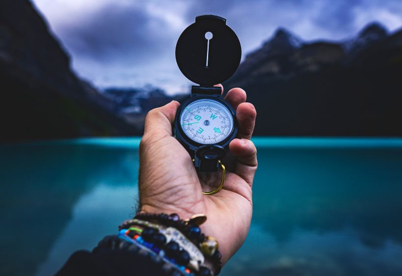 Une main tendue tenant une boussole, guidant le chemin vers un lac tranquille encadré par de majestueuses montagnes au loin.