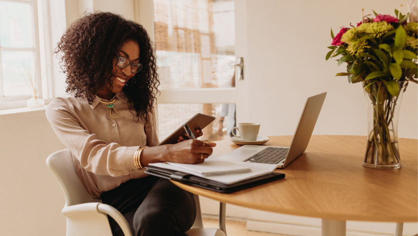 Une femme souriante est assise à une table, avec un ordinateur portable et une tasse de café devant elle. Tenant une tablette dans une main, elle prend des notes sur un bloc-notes placé à côté de son 