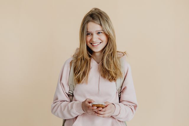 Une personne adolescente souriante aux cheveux blonds, portant un chandail rose et utilisant un téléphone intelligent à l’intérieur.