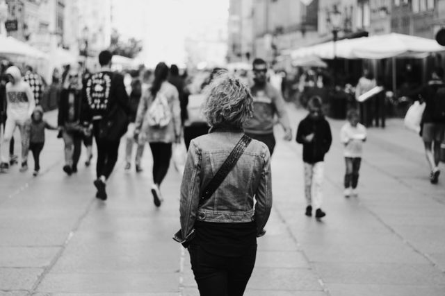 Photo en noir et blanc d’une rue urbaine achalandée où circulent des piétonnes et des piétons, mettant l’accent sur une femme vue de dos.