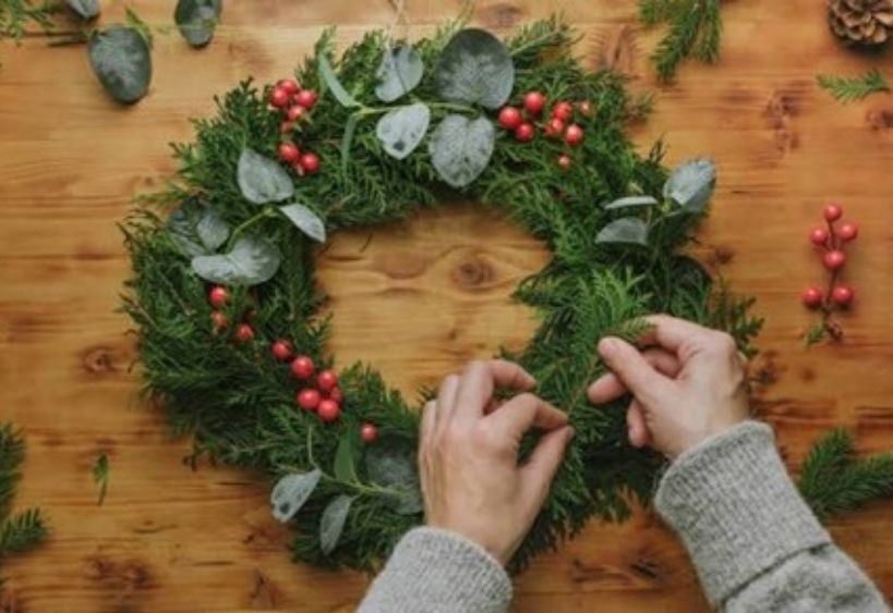 Une couronne des Fêtes composée de branches de conifères, d’eucalyptus et de baies rouges est confectionnée avec soin sur une table en bois.