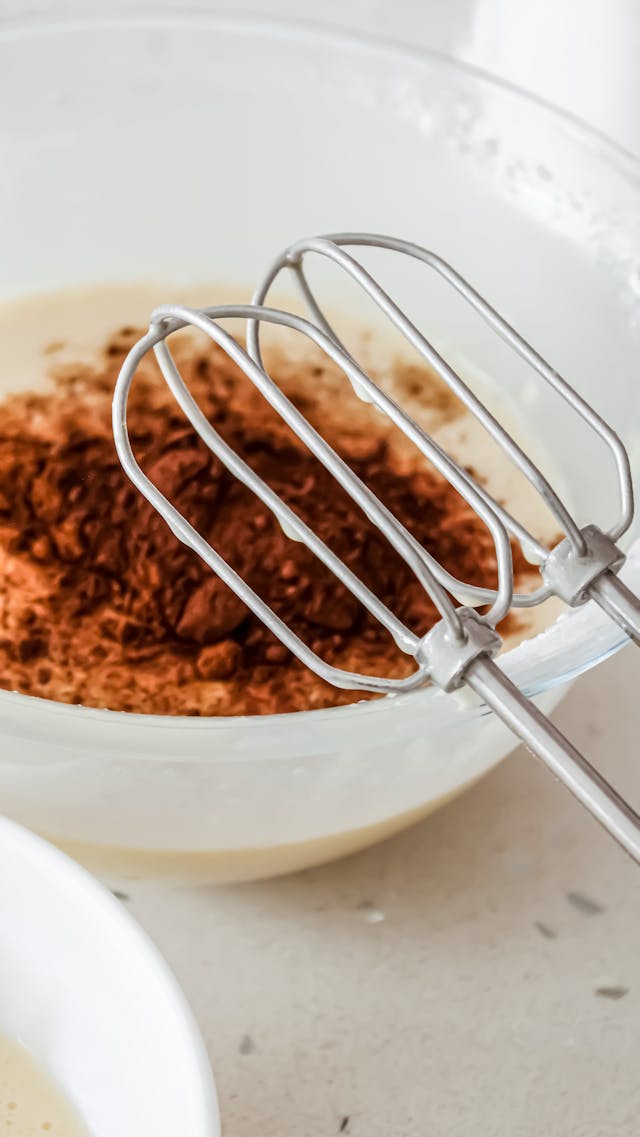 Gros plan d’un fouet à œufs et de cacao en poudre dans un bol, parfait pour un thème de pâtisserie.