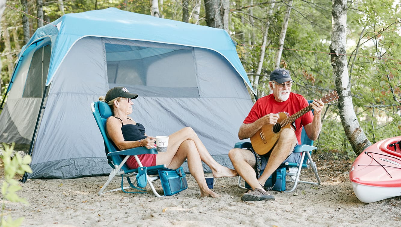 Couple en camping assis devant une tente. Homme jouant de la guitare, femme buvant du café